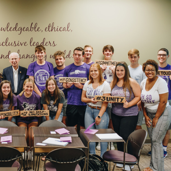 A group of students along with Bill Snyder look at the camera and hold signs with words "unity", "consistency", "commitment" and others 