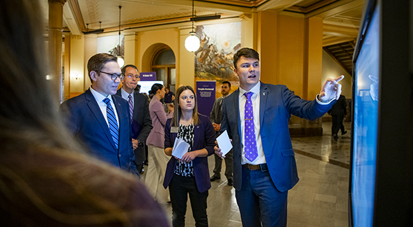 A college student in a suit presents on a portable touchscreen to legislators at a state capitol.