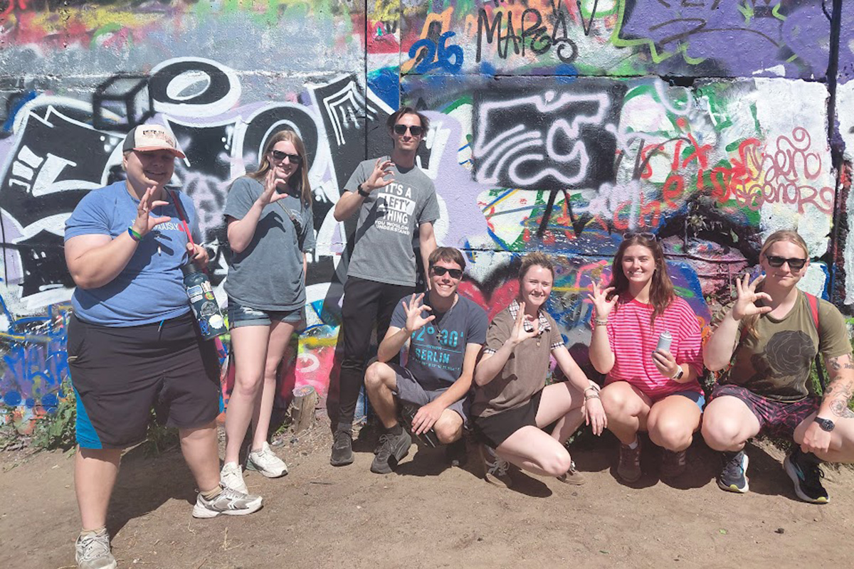 A group of college students stands in front of the Berlin Wall for a group photo. 