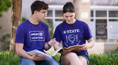 Two students sit outside of the K-State Student Union. They compare notes in their notebooks.