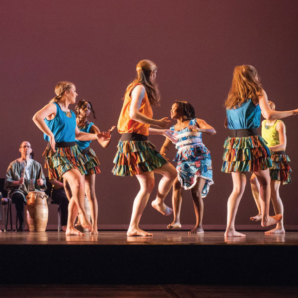 A group of dance students in costume perform in a small group on stage as a drum is played behind them.