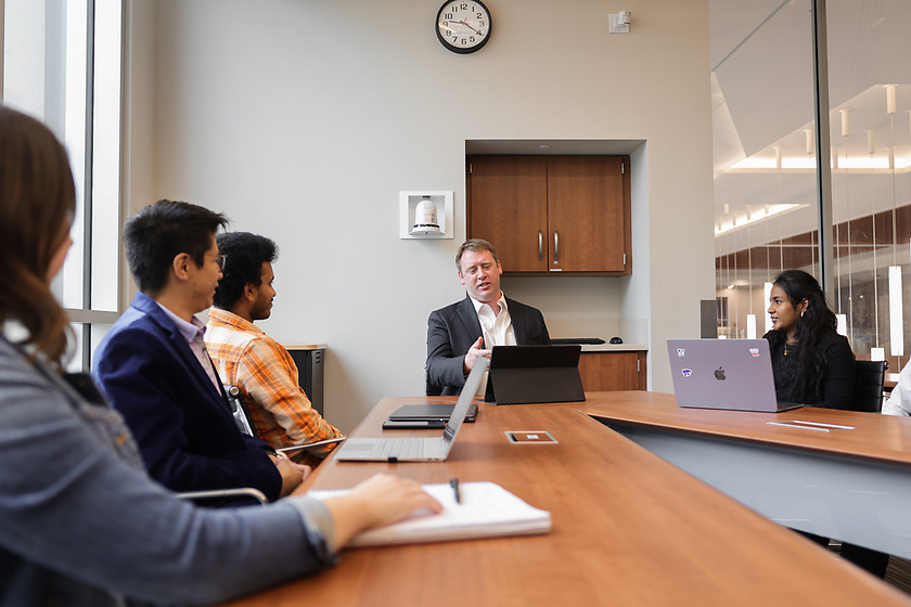 Boardroom with gentleman sitting in the center, wearing a suit and white dress shirt. A woman in all black sits to his left. Two males and a female sit to this right. 