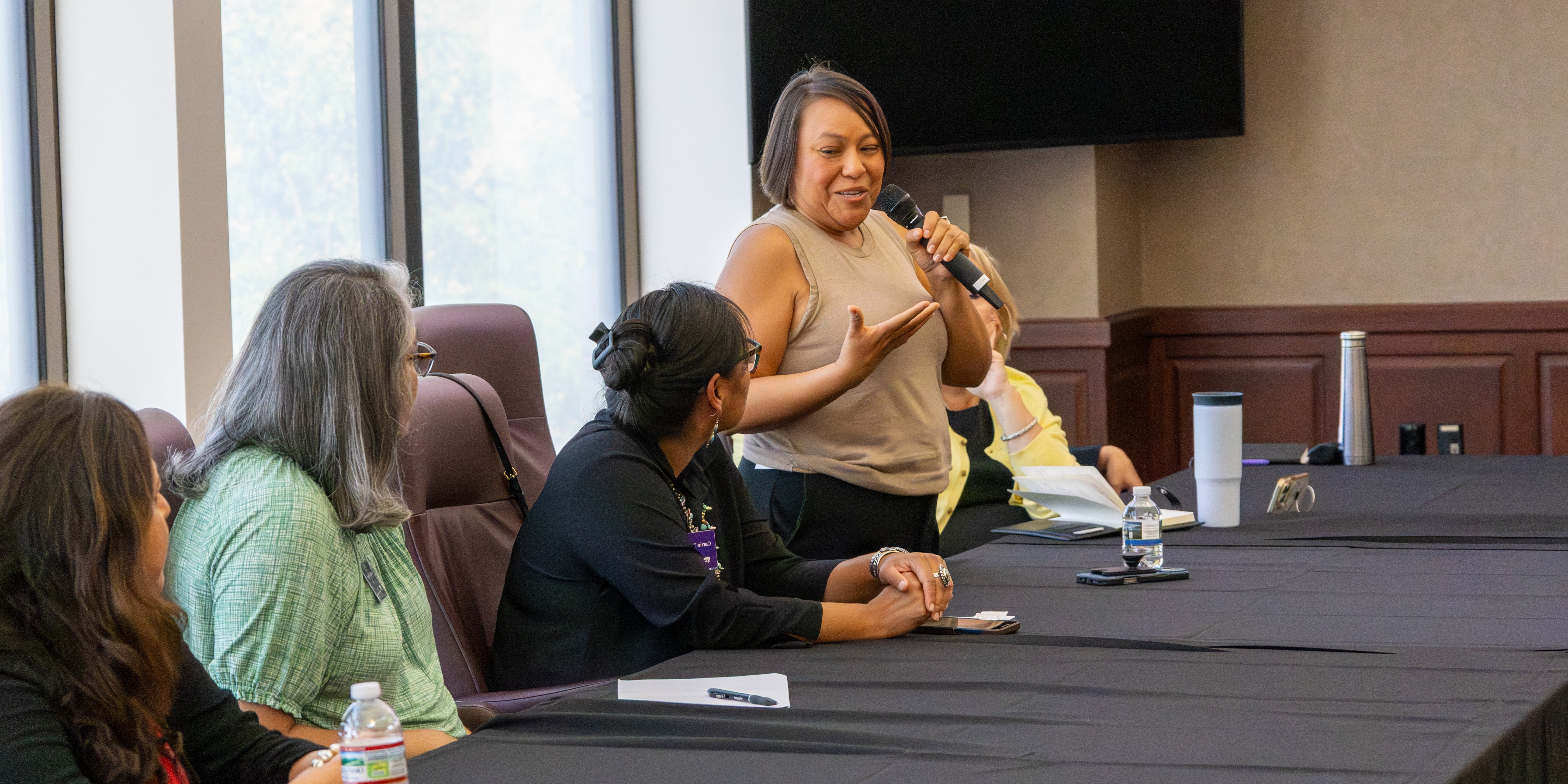 A woman stands up at a table with a microphone to speak with her colleagues.