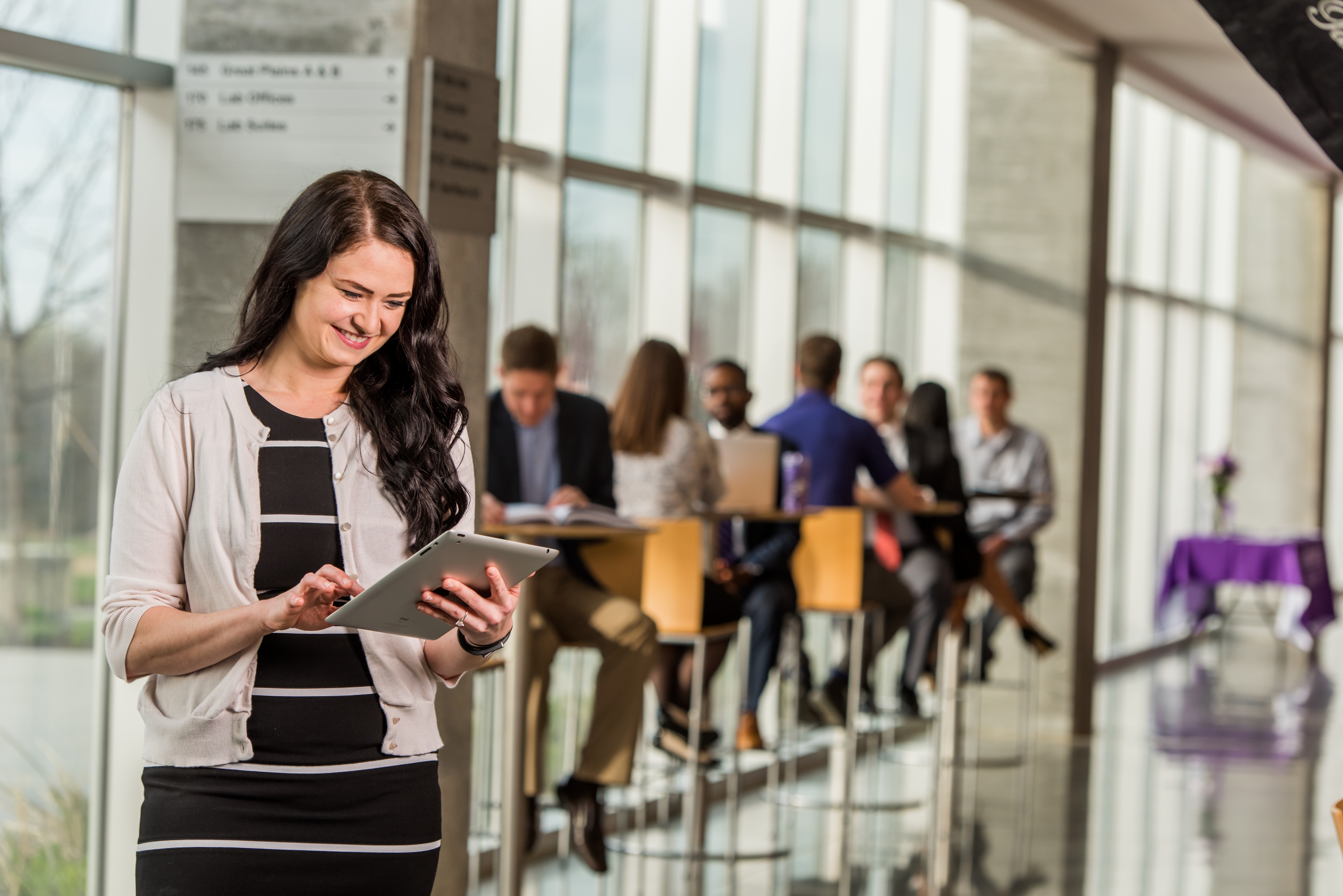 Image of business woman on a tablet in a building with a wall of windows. Other employees sit in the background at tall tables.