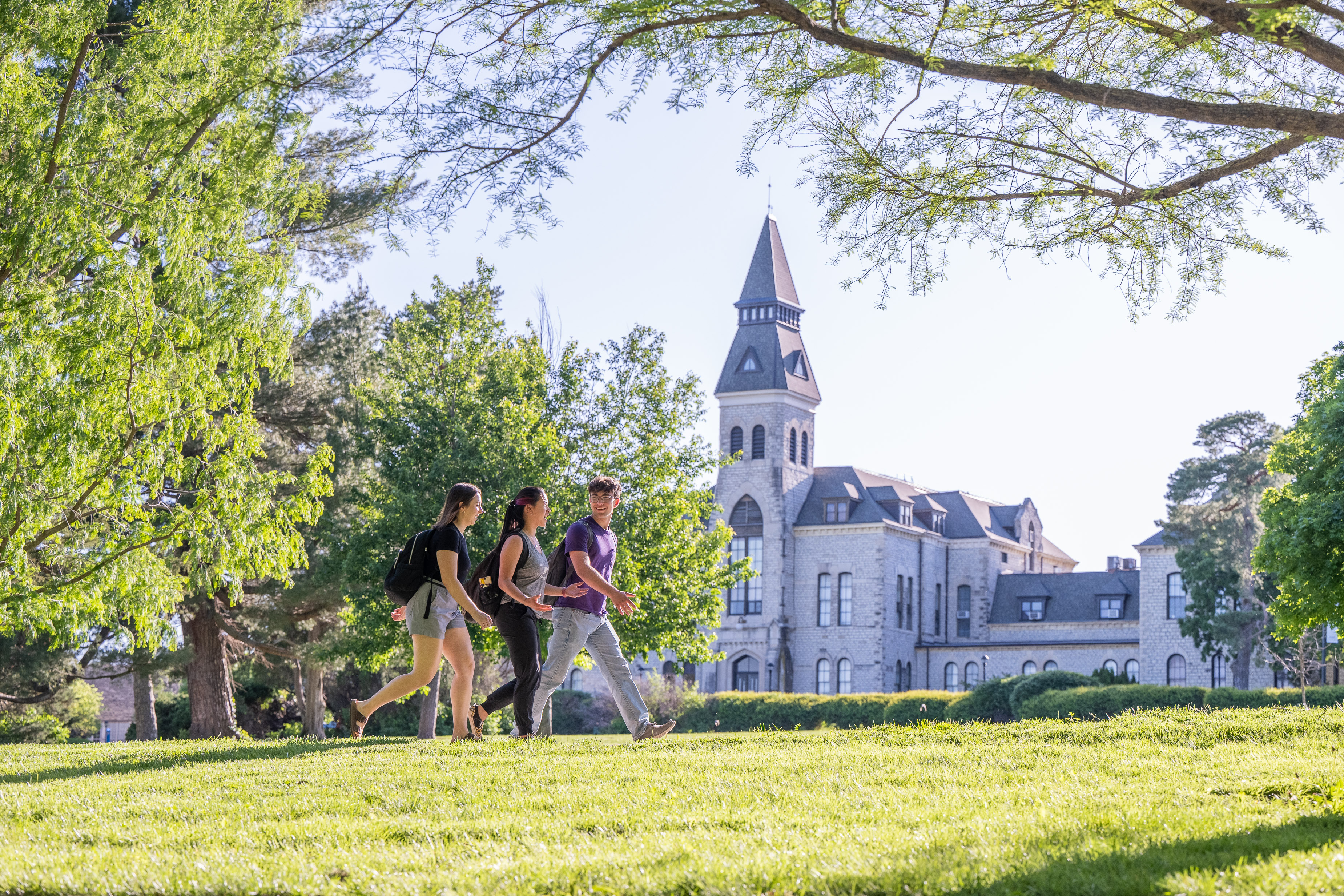 Three students walking in front of Anderson Hall with green grass and full trees. 