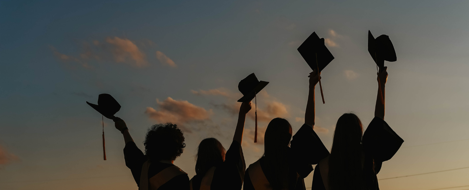 4 people wearing graduation gowns holding up their caps against a sunset