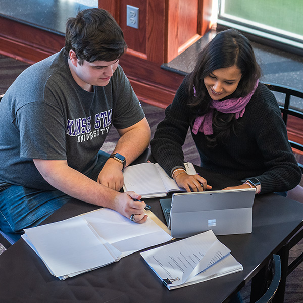 Two students study together at a table, staring at a computer with notes spread between them