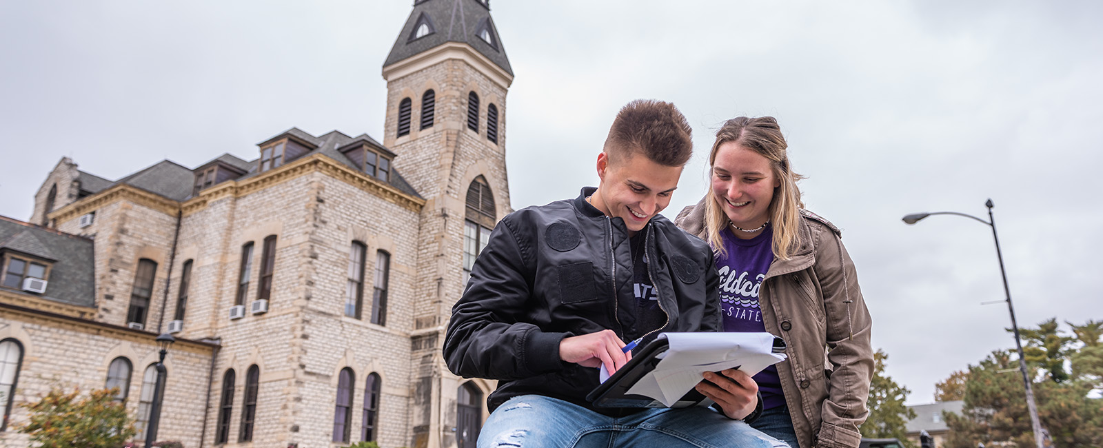 two students smiling, looking at a notebook in front of them with sky and anderson hall behind them