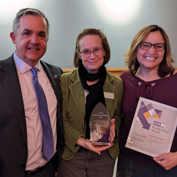 Three faculty members standing side by side and smiling. Two are holding up awards.