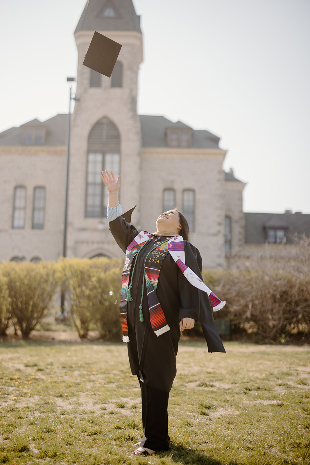A first-generation graduate tosses her cap into the air in front of Anderson Hall. She wears graduation regalia, including a green cord signifying her first-gen status