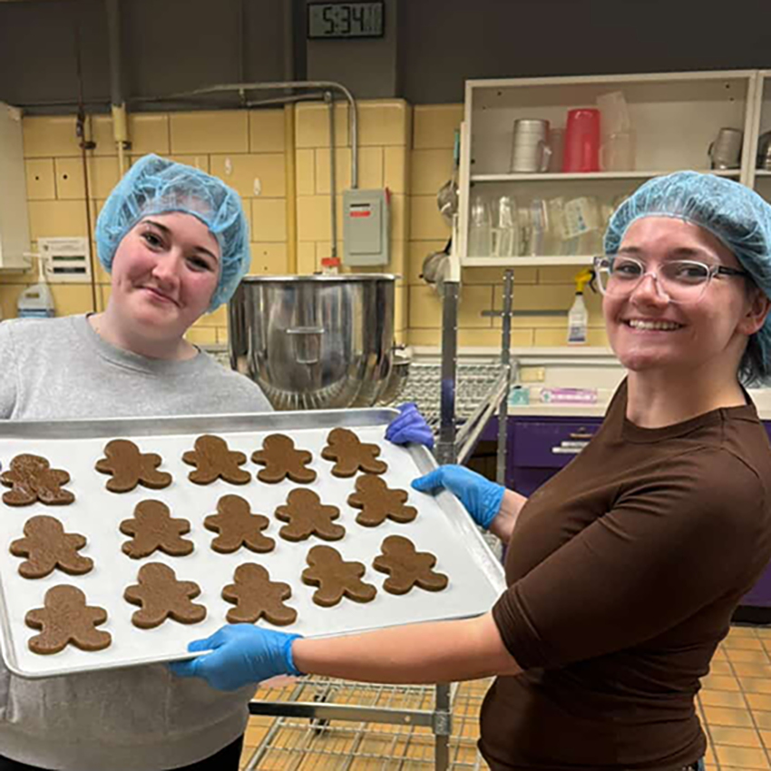 Two students wearing blue hairnets hold a silver tray of gingerbread person cookies in an industrial kitchen.