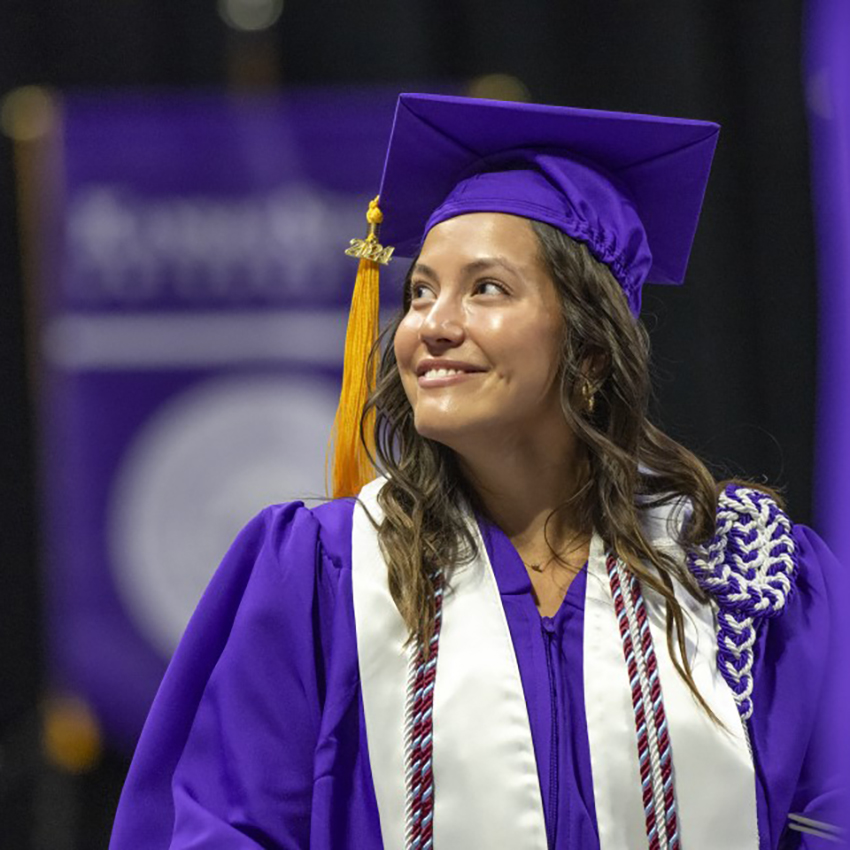 A graduate with long brown hair wearing a purple cap and gown with a gold tassel looks over her shoulder and smiles.