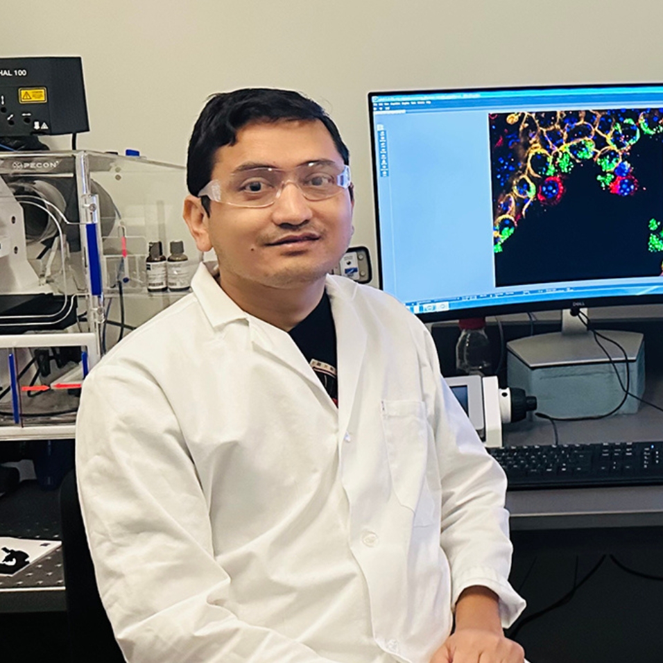 A doctoral student in a lab coat sits at a computer station and poses for a portrait.