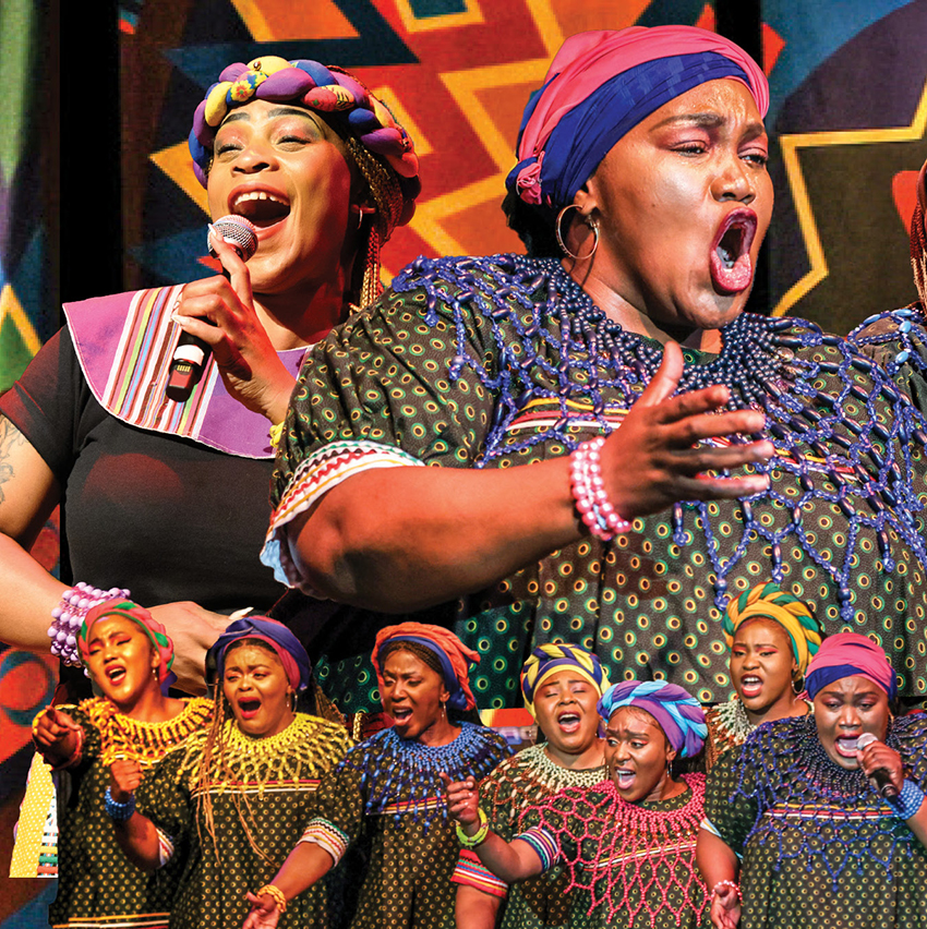 A group of women in African cultural attire sing on a stage.