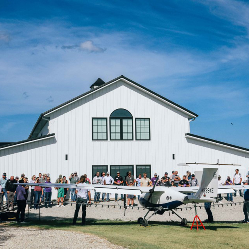 A crowd in front of a wide, white barn watches a man present in front of a large, unmanned aerial plane.