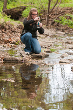 A woman in jeans and a black jacket wears safety glasses as she kneels on the rocks by a smooth stream and looks at a water sample in a glass jar.