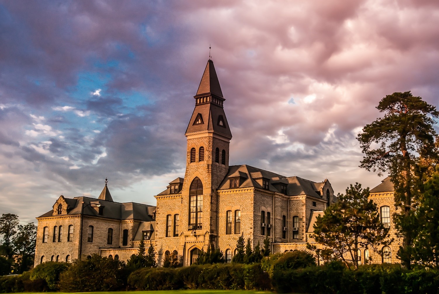Anderson Hall, K-States administration building, is made of limestone and has a bell tower. A landscape of green bushes and trees frames the building while clouds dot a blue sky behind it.