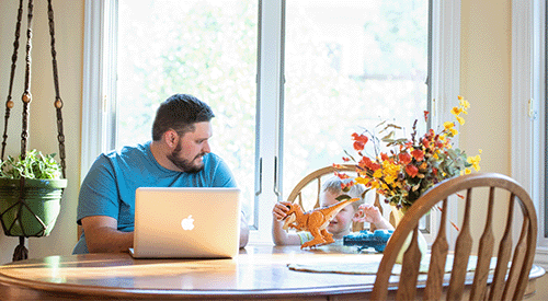 A man sits at his kitchen table, his child next to him, as he completes his education online.