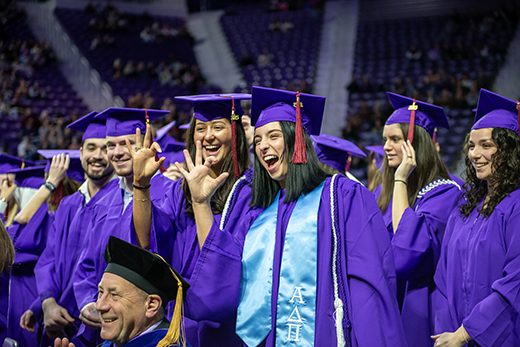 Several students in purple graduation caps and gowns flash the W-C hand symbol on the floor of Bramlage Coliseum.