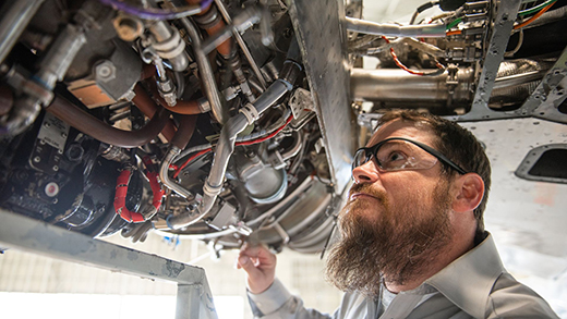 A bearded student wearing goggles pokes through the machinery in an open aircraft panel.