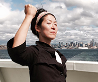 Standing on a boat in a harbor with what appears to be the New York City headline in the background, Susie Lee stands and looks at the camera. Her right fist is raised in the air and her hair is pulled back behind a white floral bandana.