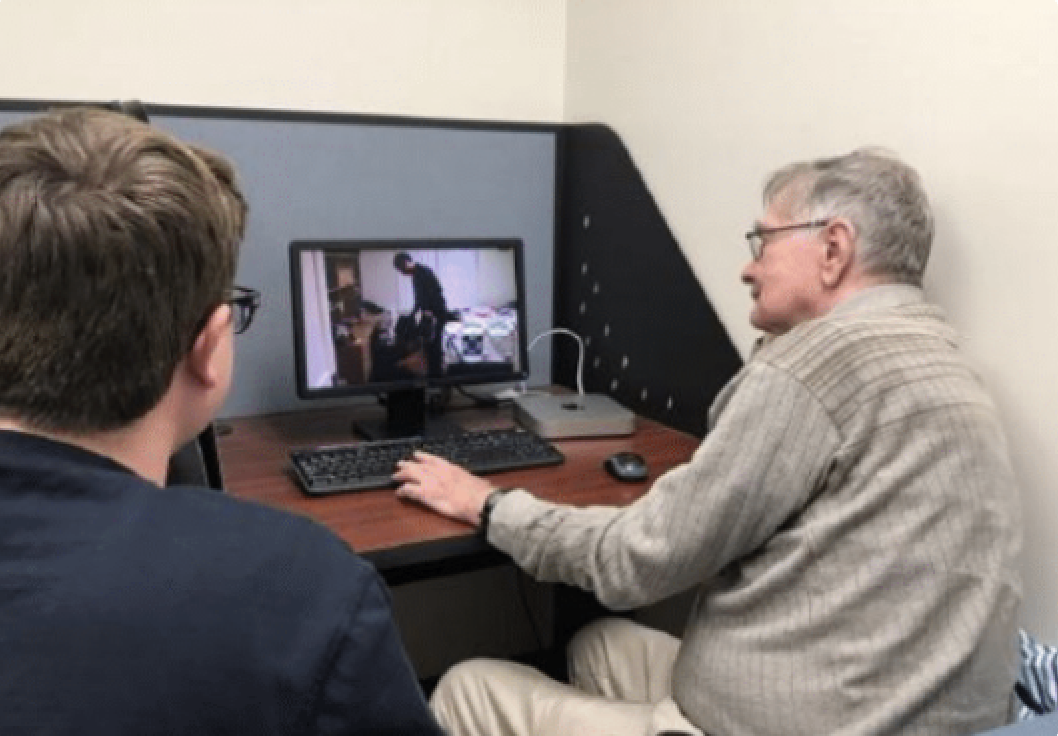 An older adult sat at a research computer, watching a video on screen. A researcher sits to the left, watching the study