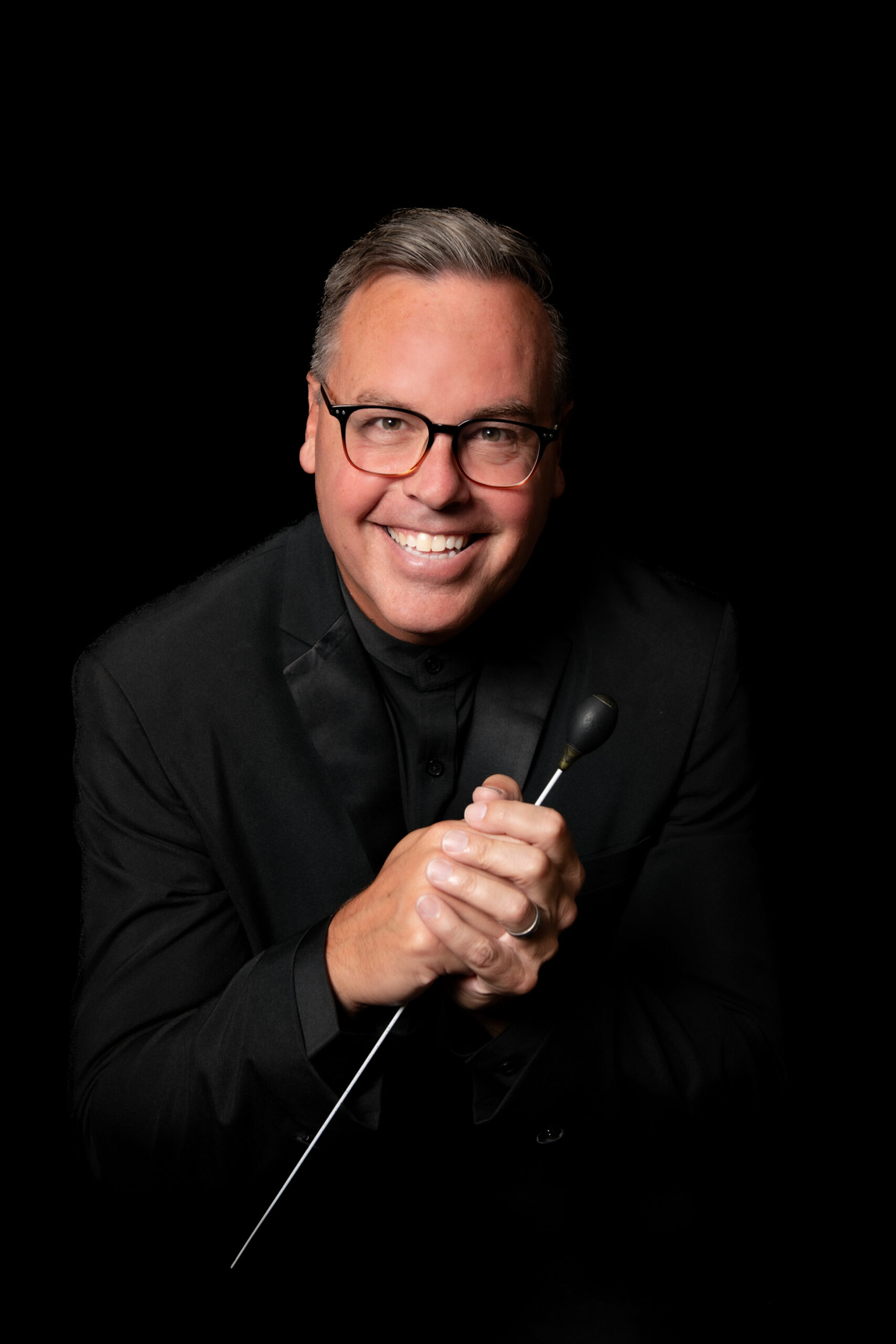 A man in a black suit holding a baton in front of a black background.