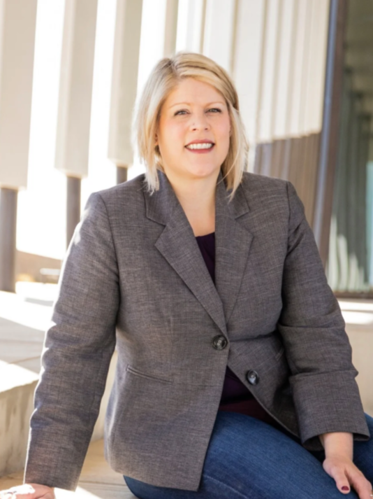 A woman in a grey blazer and jeans sits backlit by a window with vertical panes.