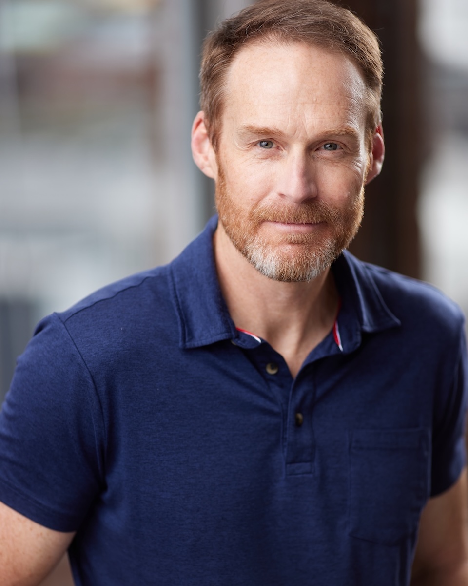A bearded man (Dwight Tolar) stands in a blue shirt in front of an unfocused backgroud.