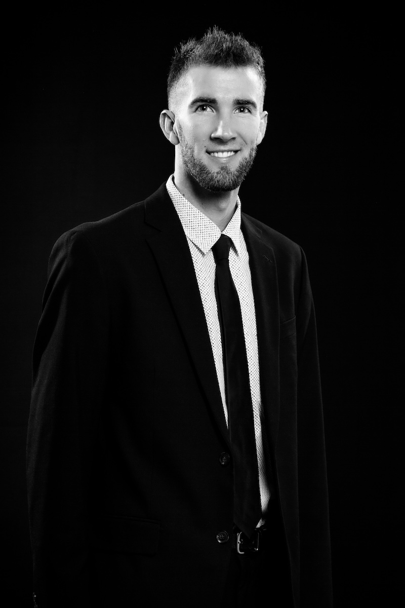 a man with a beard and dark hair in a black suit in front of a black background