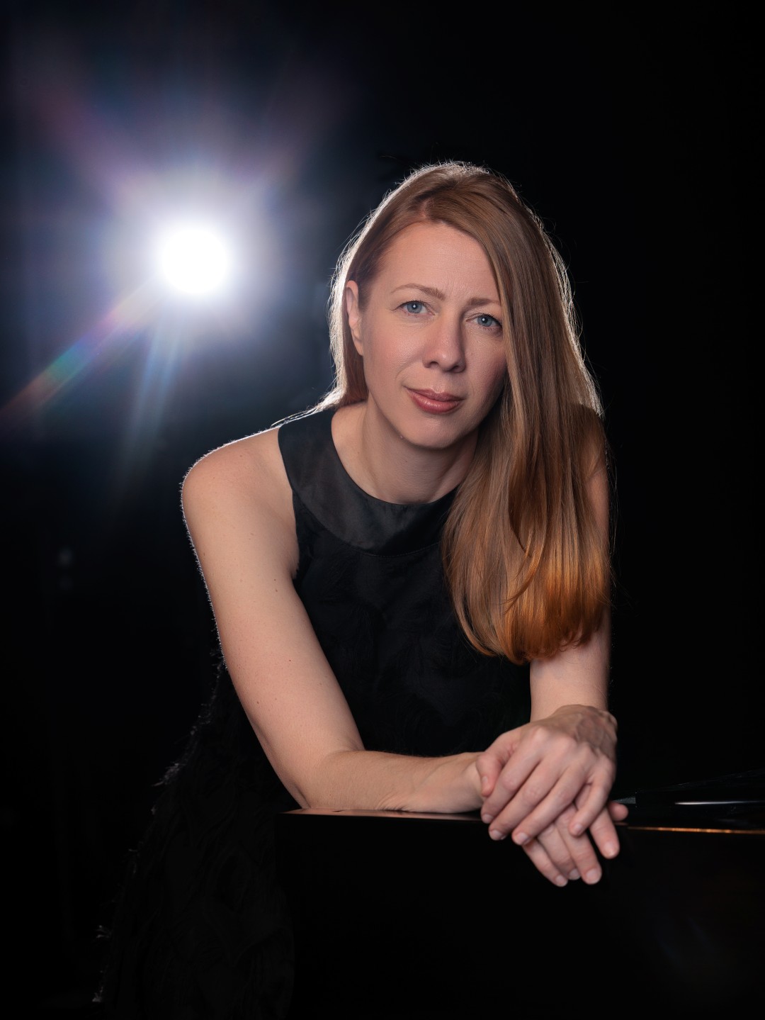 A woman leans on the top of a piano in a black dress with a spotlight shining from behind