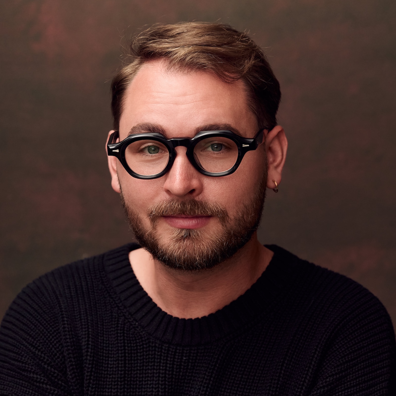 a man with a beard and glasses wearing a black shirt in front of a brown background