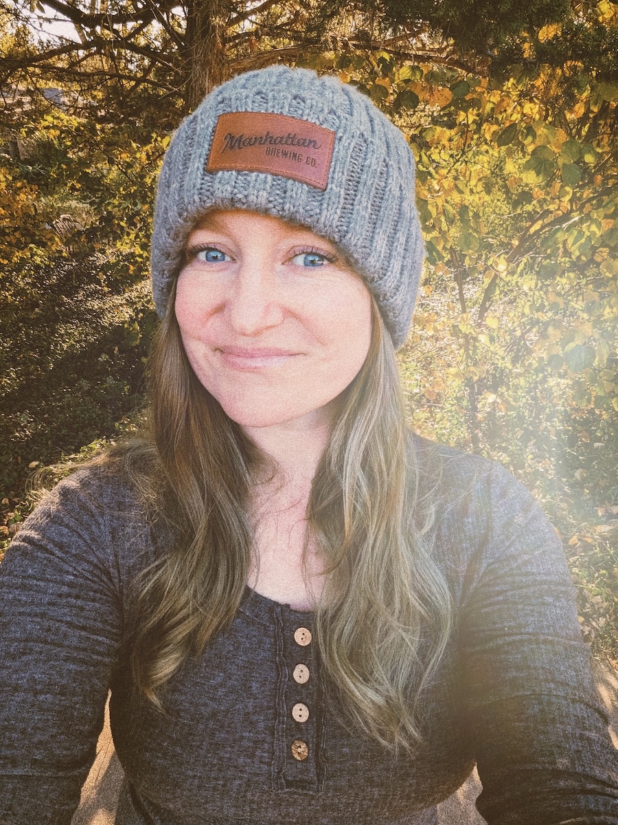 a woman in a grey stocking cap and grey shirt stands in front of a group of trees