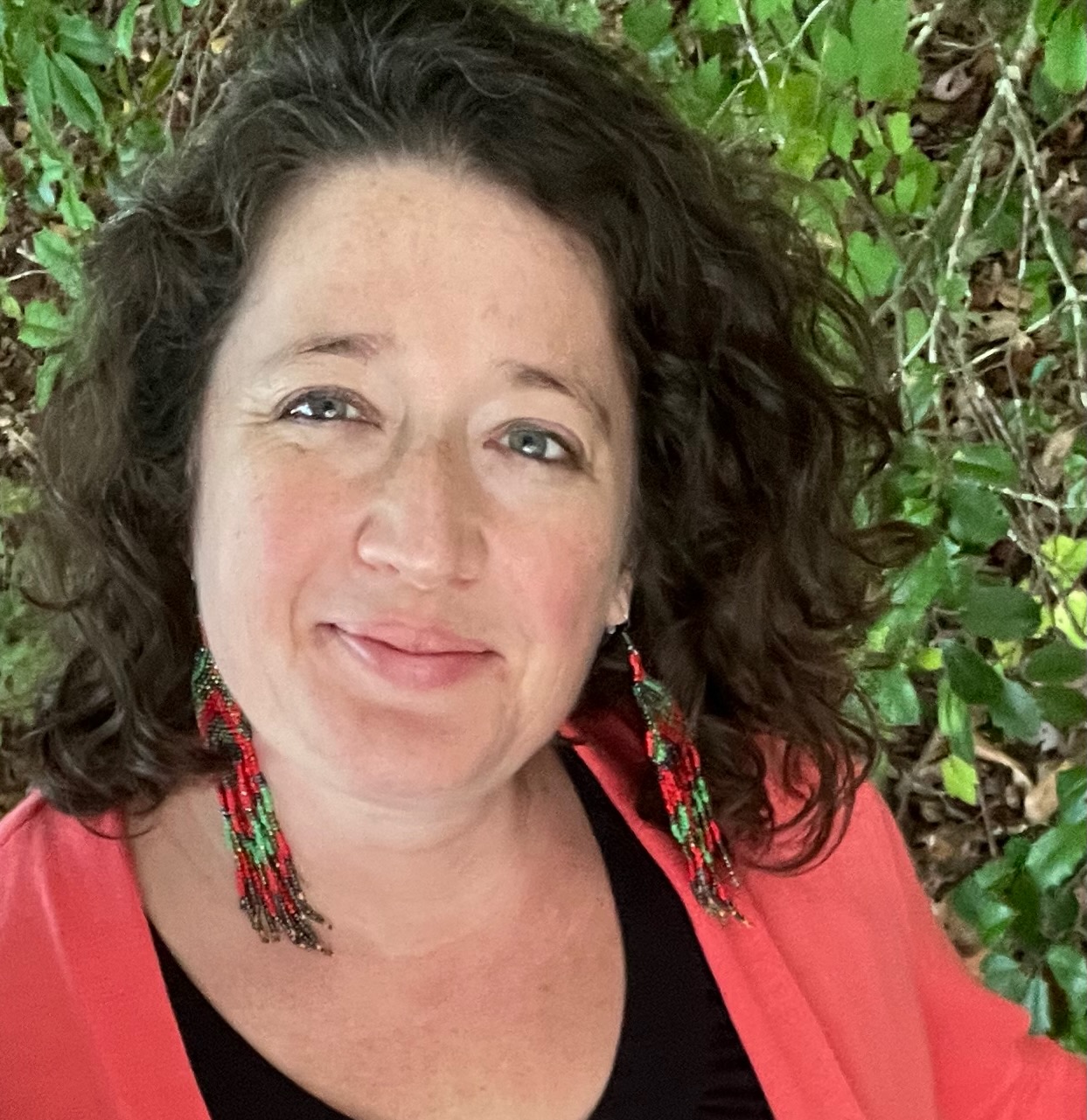 A woman with dark hair and a coral top stands in front of vegetation