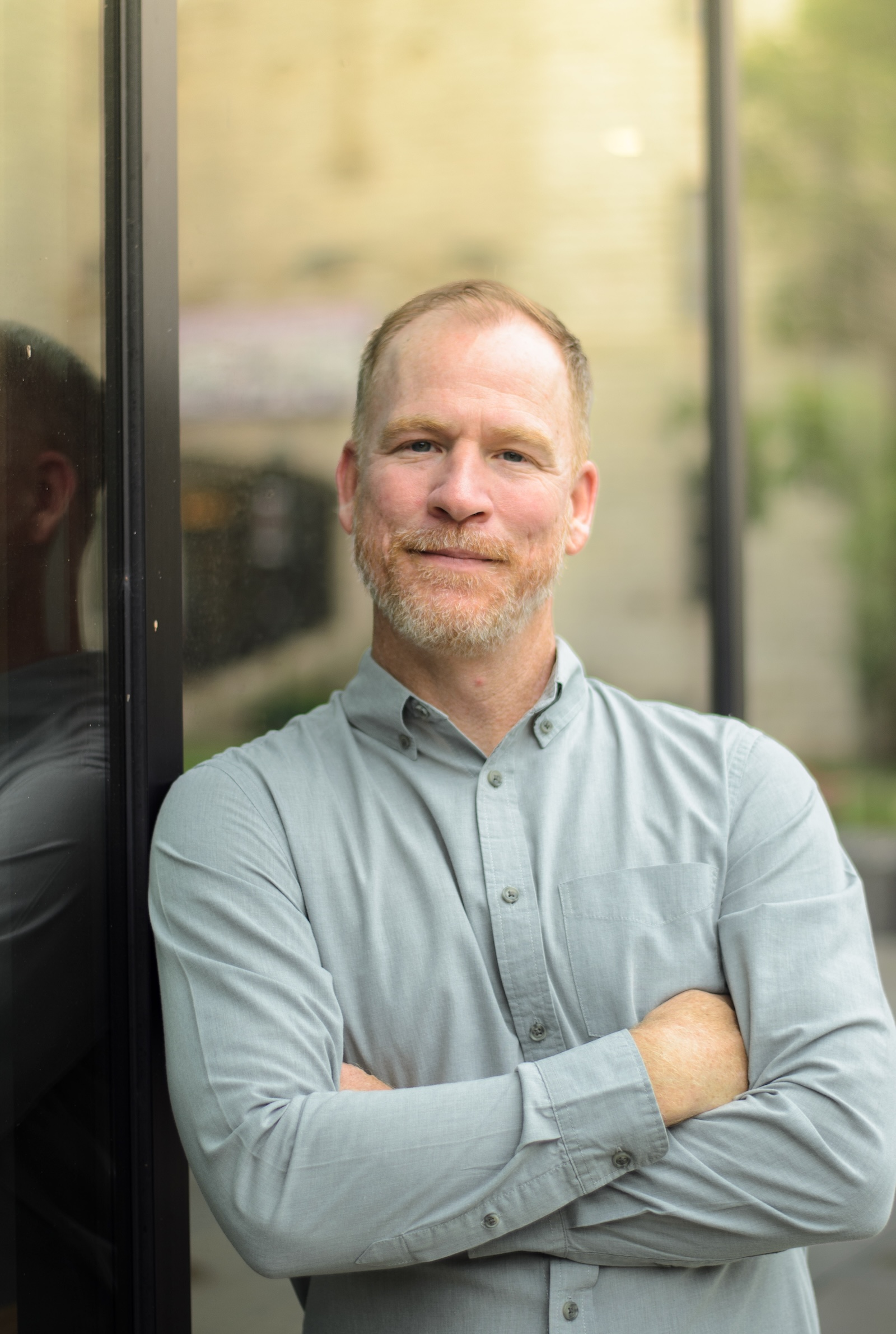 a man with a bread in a grey shirt leans against a window