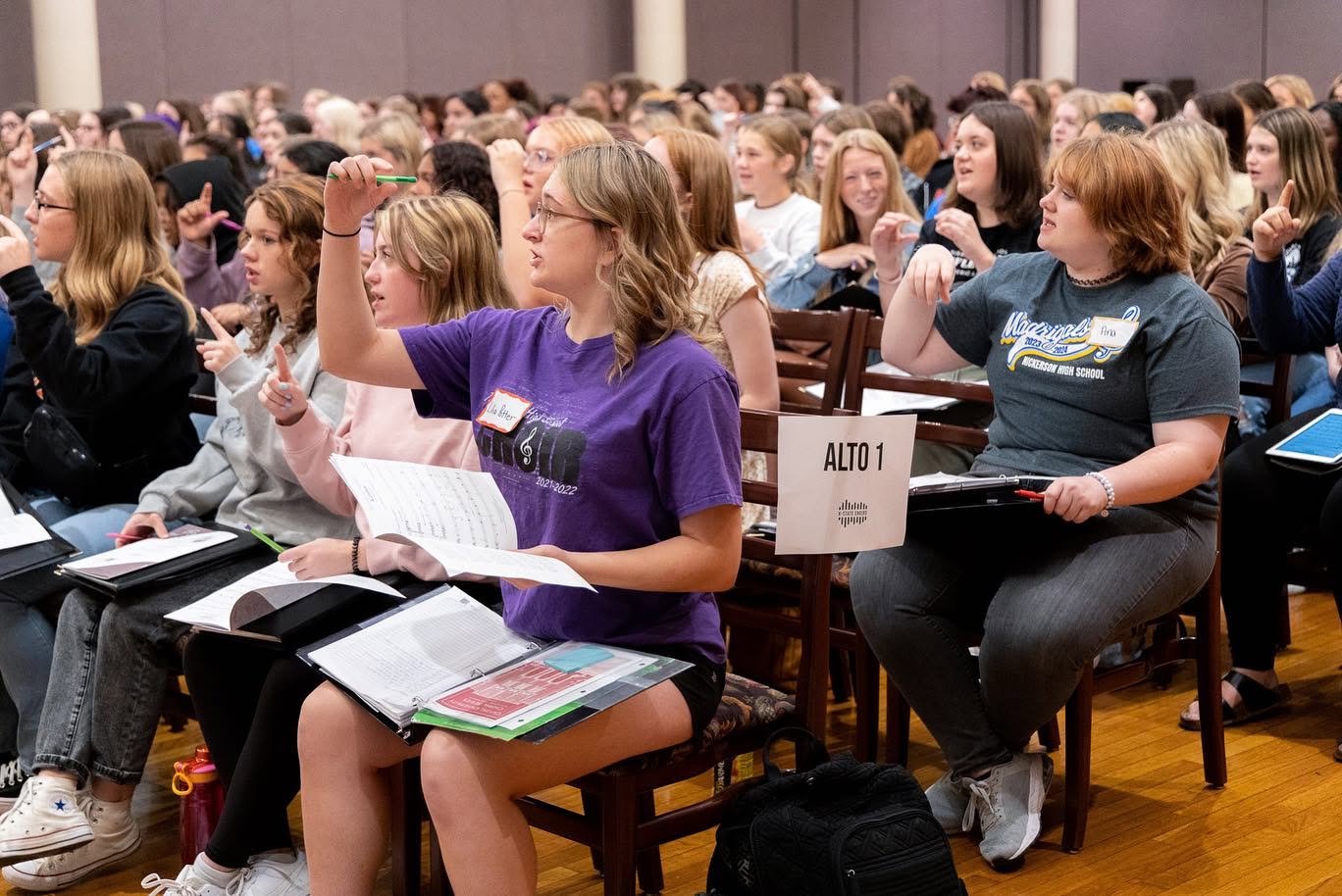 female student sit in chairs with their hands raised with sheet music on their laps.