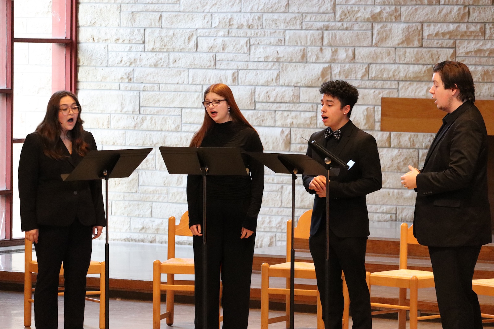 Four singers in black, stand behind music stands.