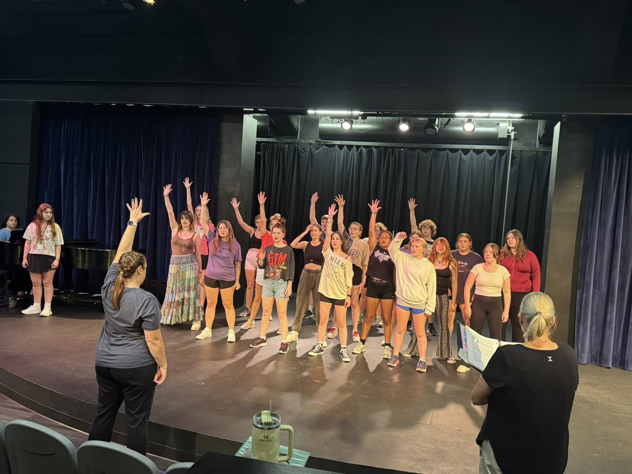 a group of performers stand in rehearsal on a stage with a single arm stretched over their head as two directors look on teaching them the movement.