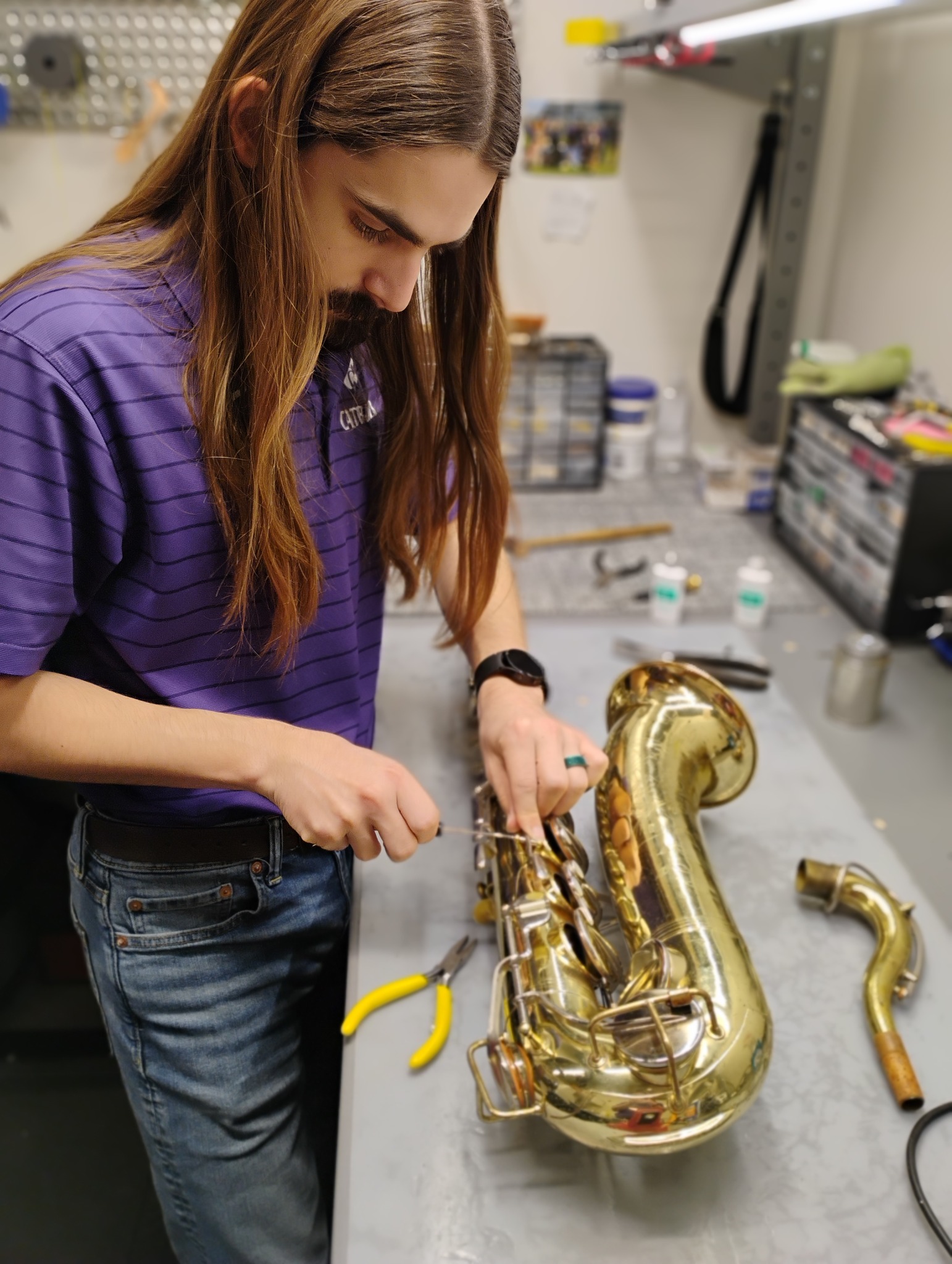 man with a beard in a purple polo shirt repairs a saxophone