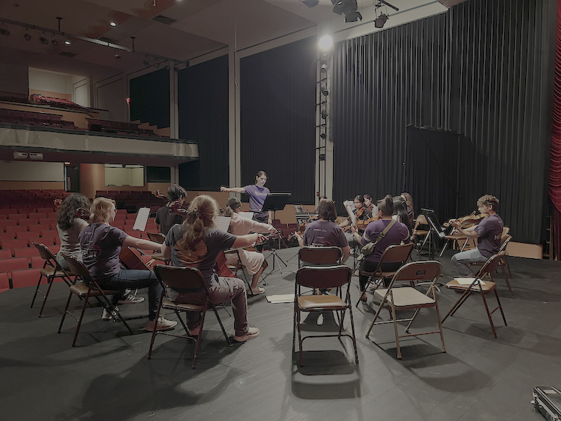 a women in a purple K-State t-shirt conducts a group of young string players on a stage.