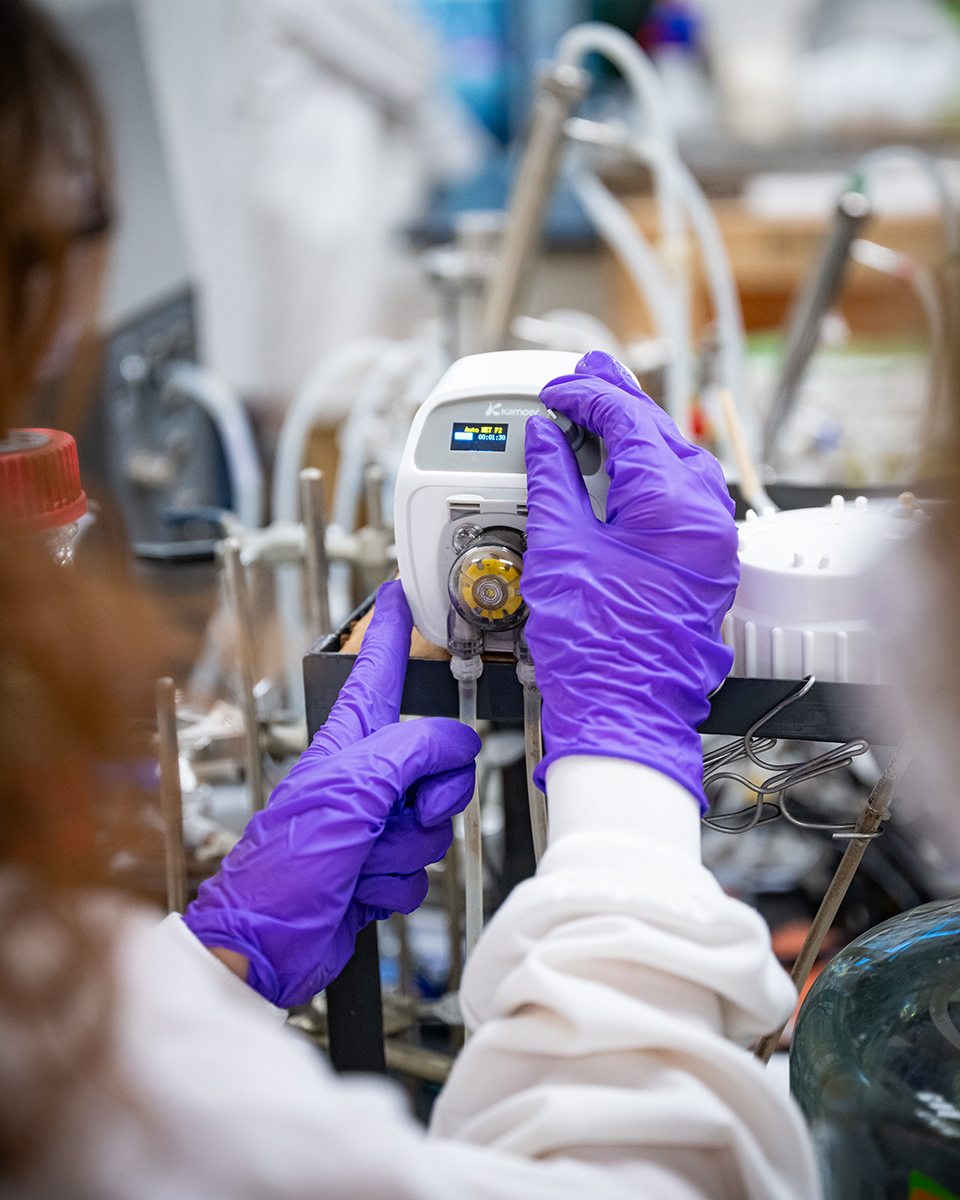 A close-up photo from behind shows someone using purple gloves to manipulate a piece of research equipment in a lab.