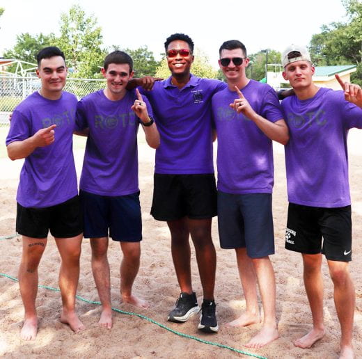 Army ROTC men in purple ROTC t-shirts, smiling and standing barefoot in sand making "#1" hand gesture.