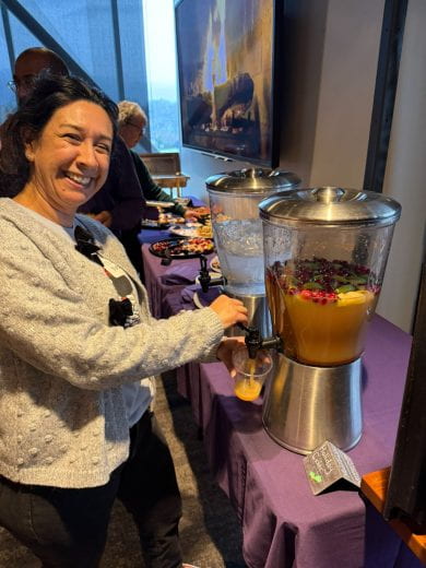 smiling guest at food table pours drink from canister containing cider and multicolored fruit