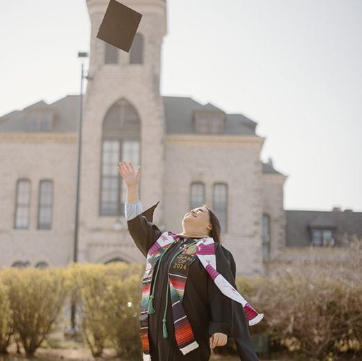 photo of female in graduation regalia in front of Anderson Hall throwing cap up in air