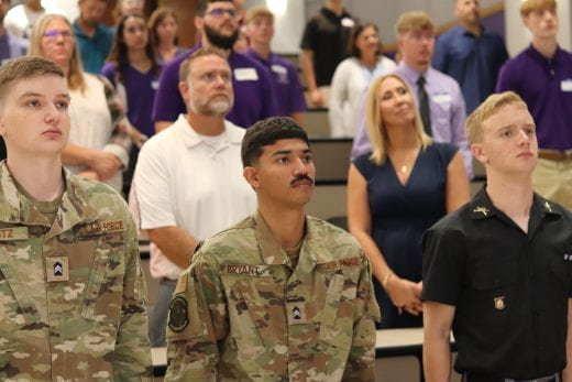 3 cadets stand at attention in foreground with other audience members in background
