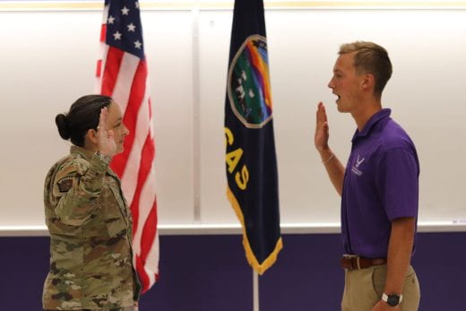 A camouflage-uniformed woman and young man in purple polo shirt face each other with a hand each raised as he appears to be swearing an oath