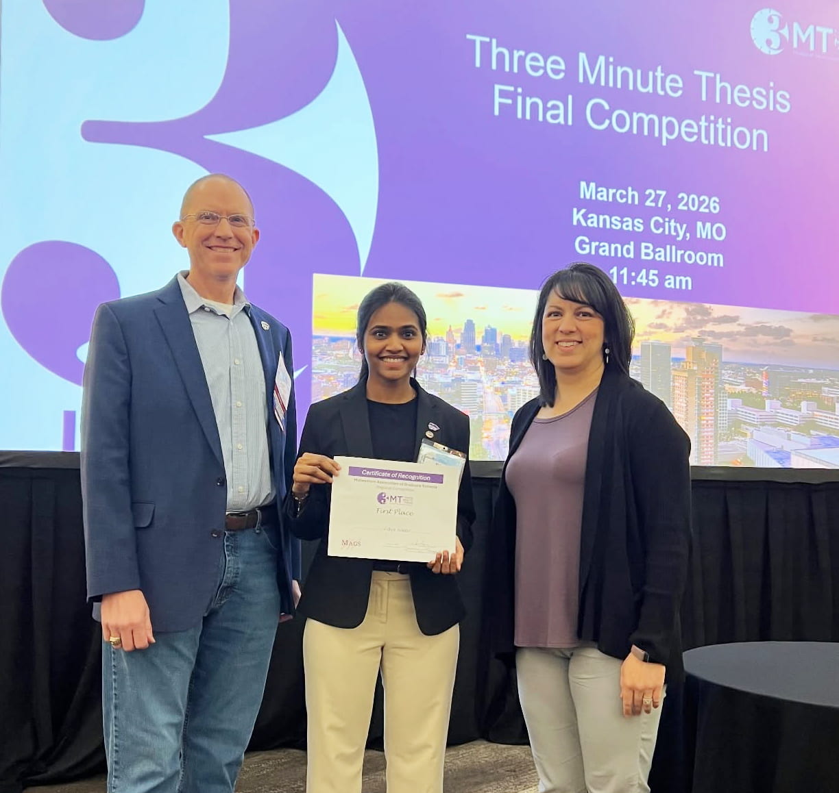 Vidya Nadar, posed with two others, holds winner certificate with screen in background displaying 3MT event info