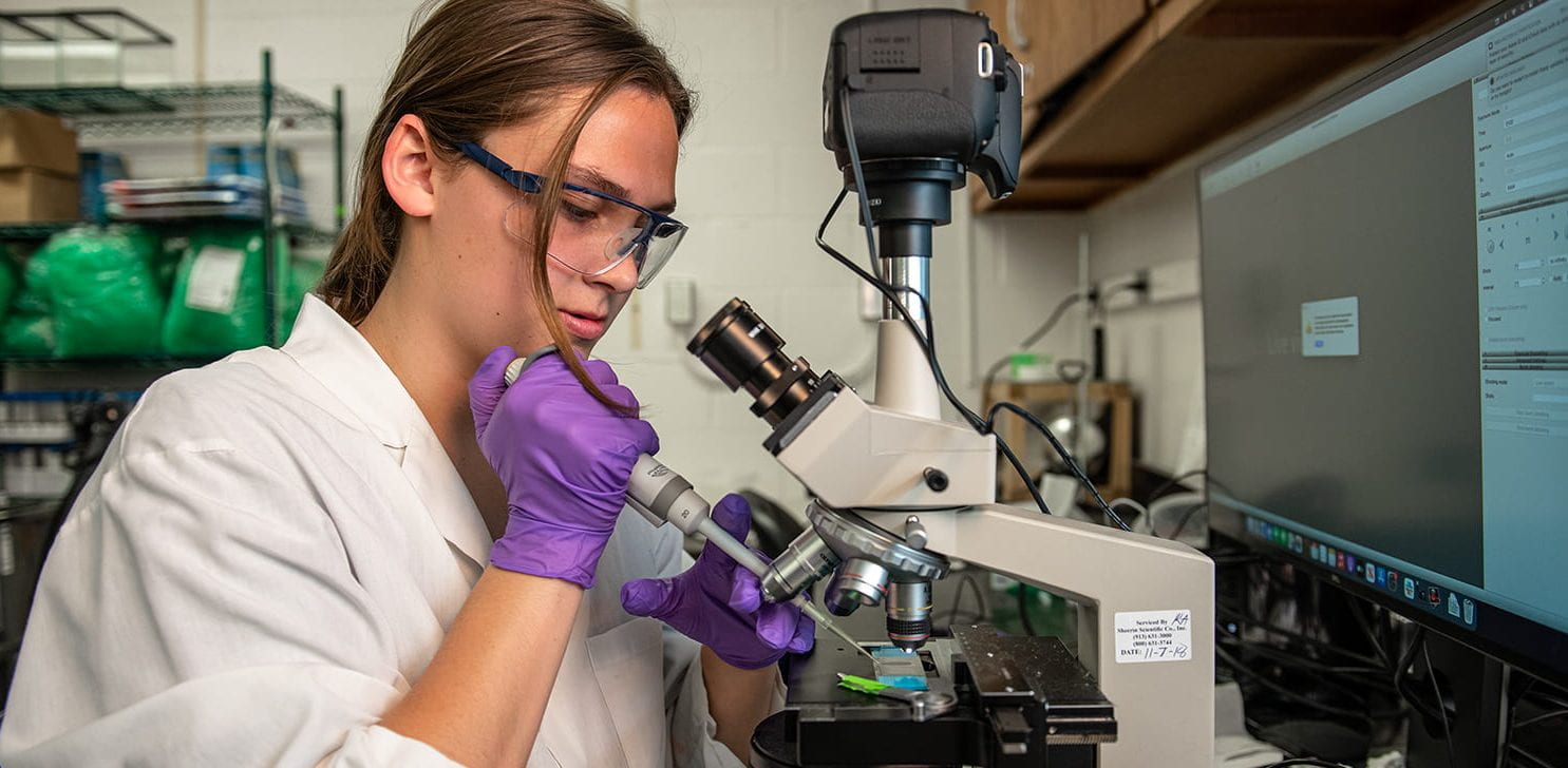 student pipetting at microscope in white lab coat and purple gloves