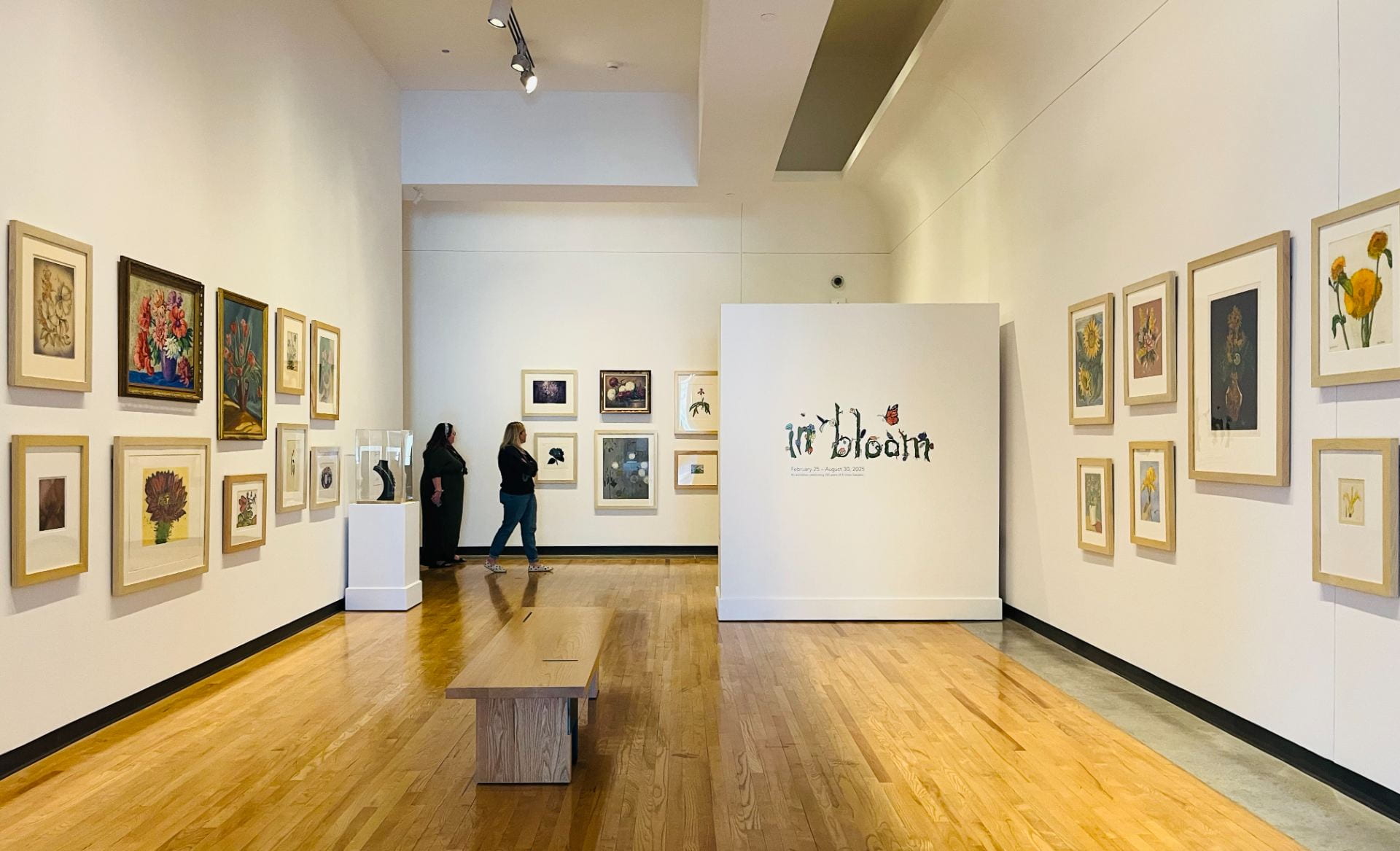 "In Bloom" exhibition gallery with floral artworks displayed on the walls. There is a title wall with "In Blom" text designed using various floral elements like flowers, leaves, birds etc. 