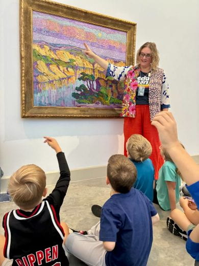 Art teacher and children discussing a painting on the wall. Teacher pointing at the painting and student raise their hands in response.